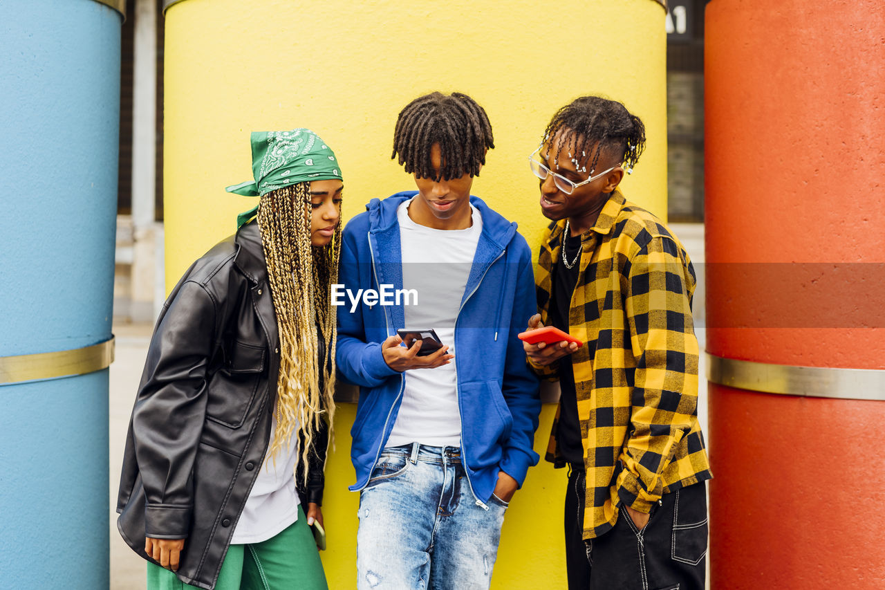 Young man sharing smart phone with friends in front of yellow pipe