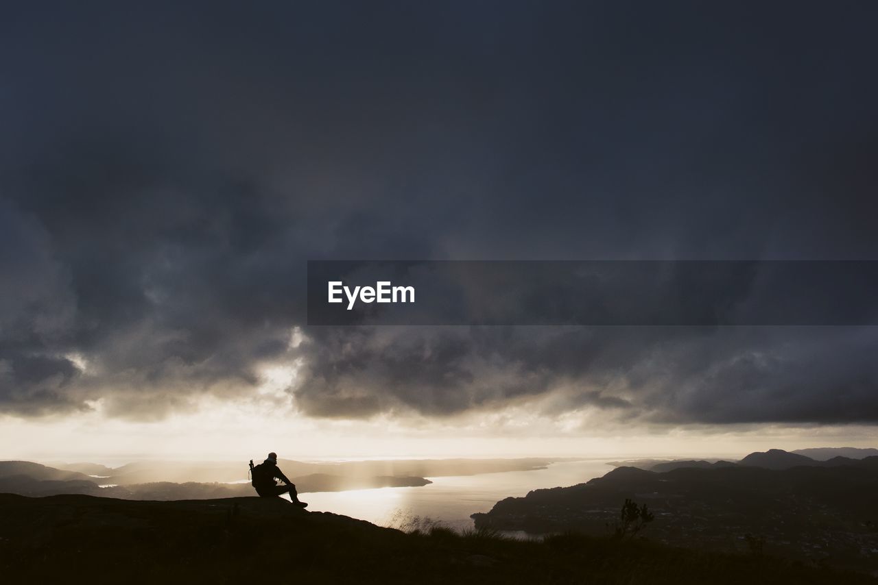 Silhouette man sitting on mountain against stormy clouds during sunset