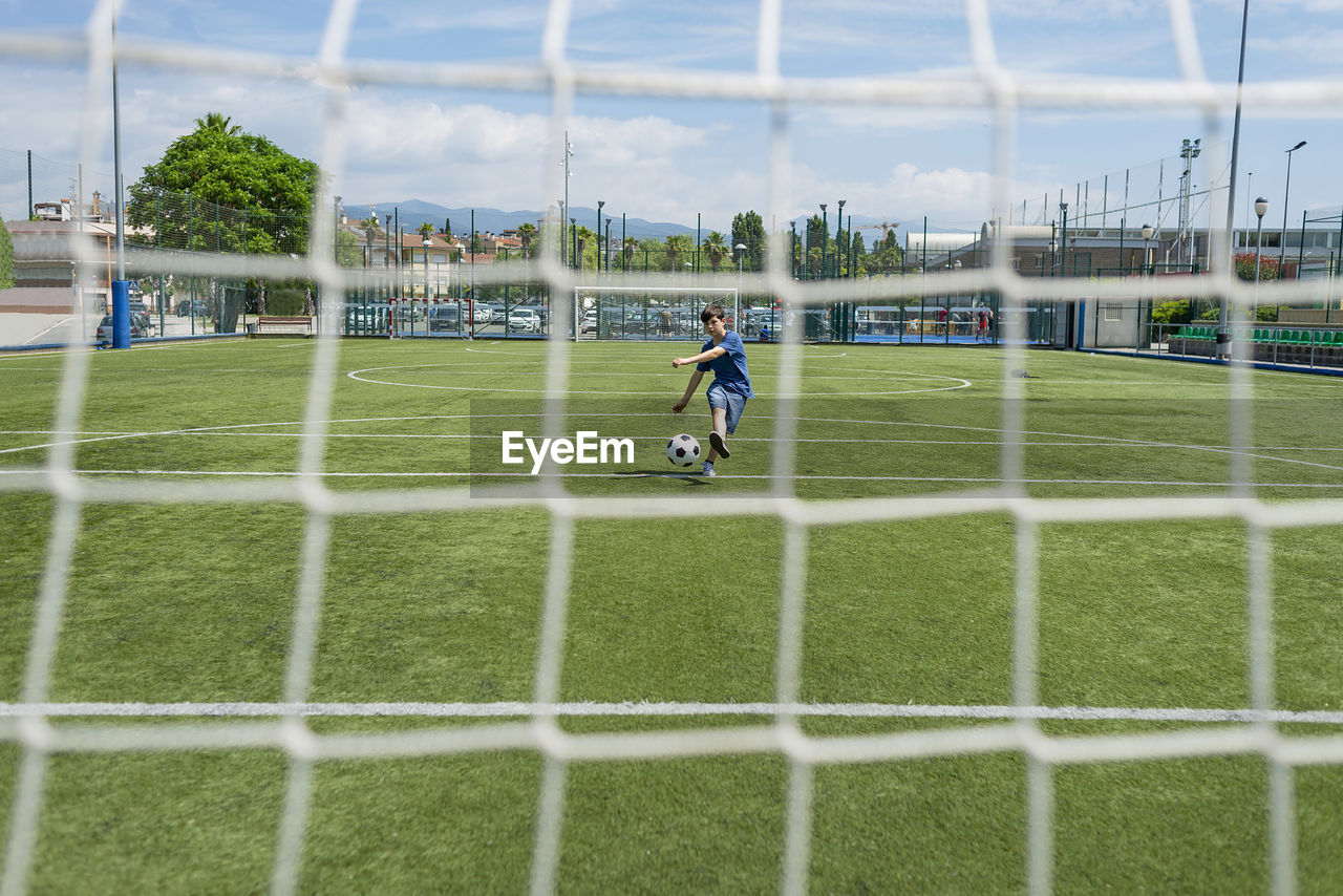 Boy kicking soccer ball against sky seen through net
