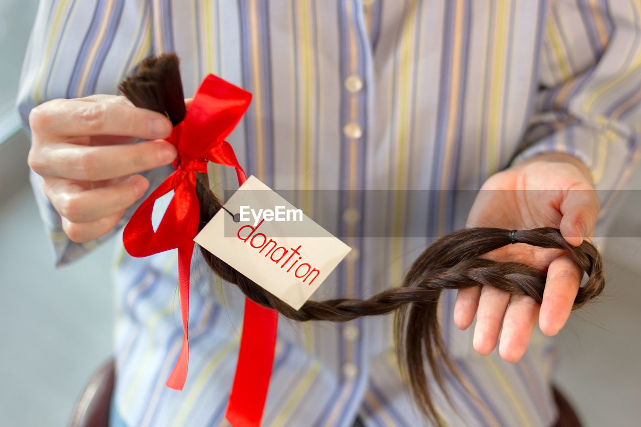 Long cut braid with tag and red ribbon bow in woman hands, donation of haircut, faceless concept
