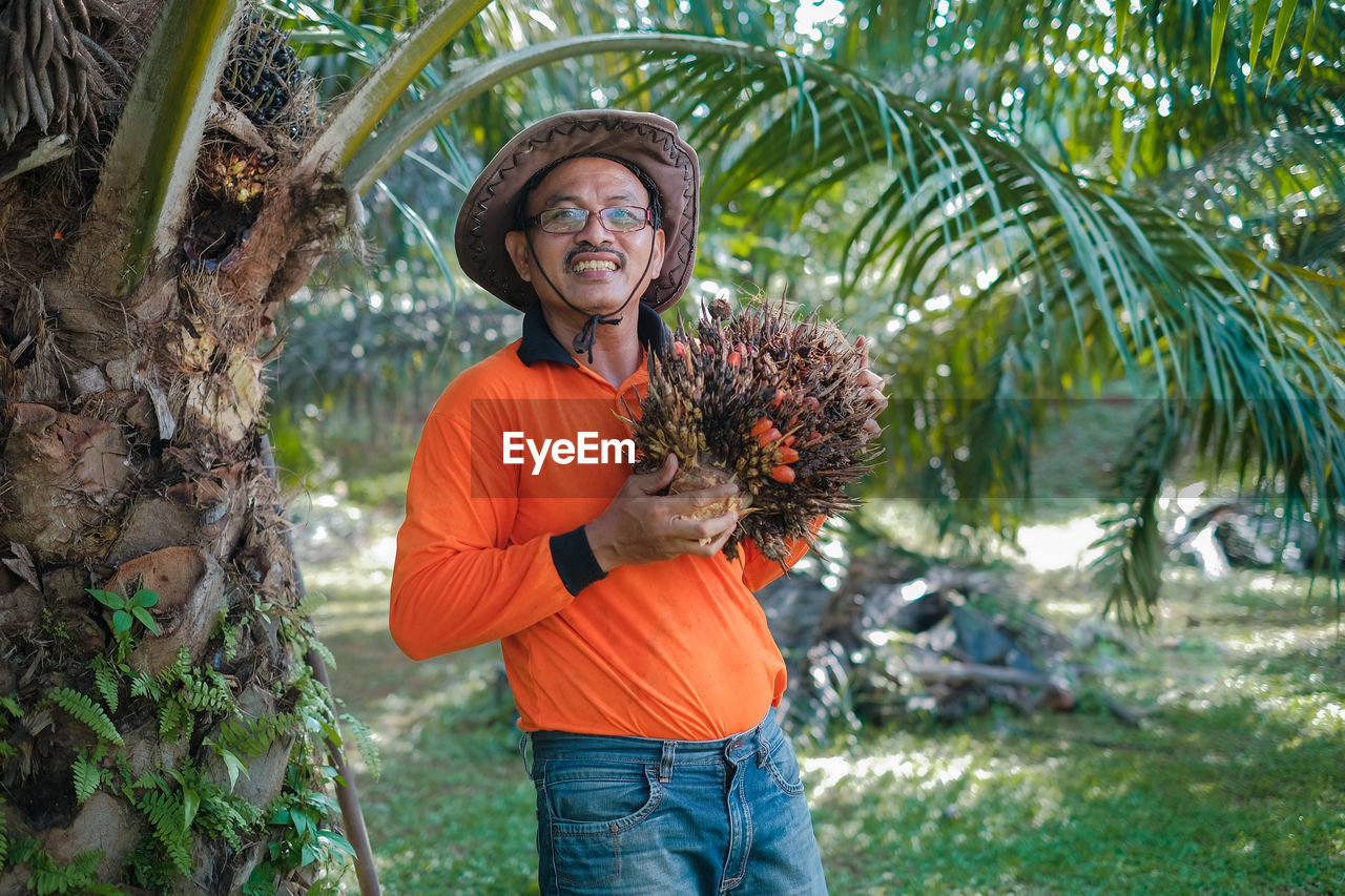 Portrait of man wearing hat holding palm fruit standing by tree