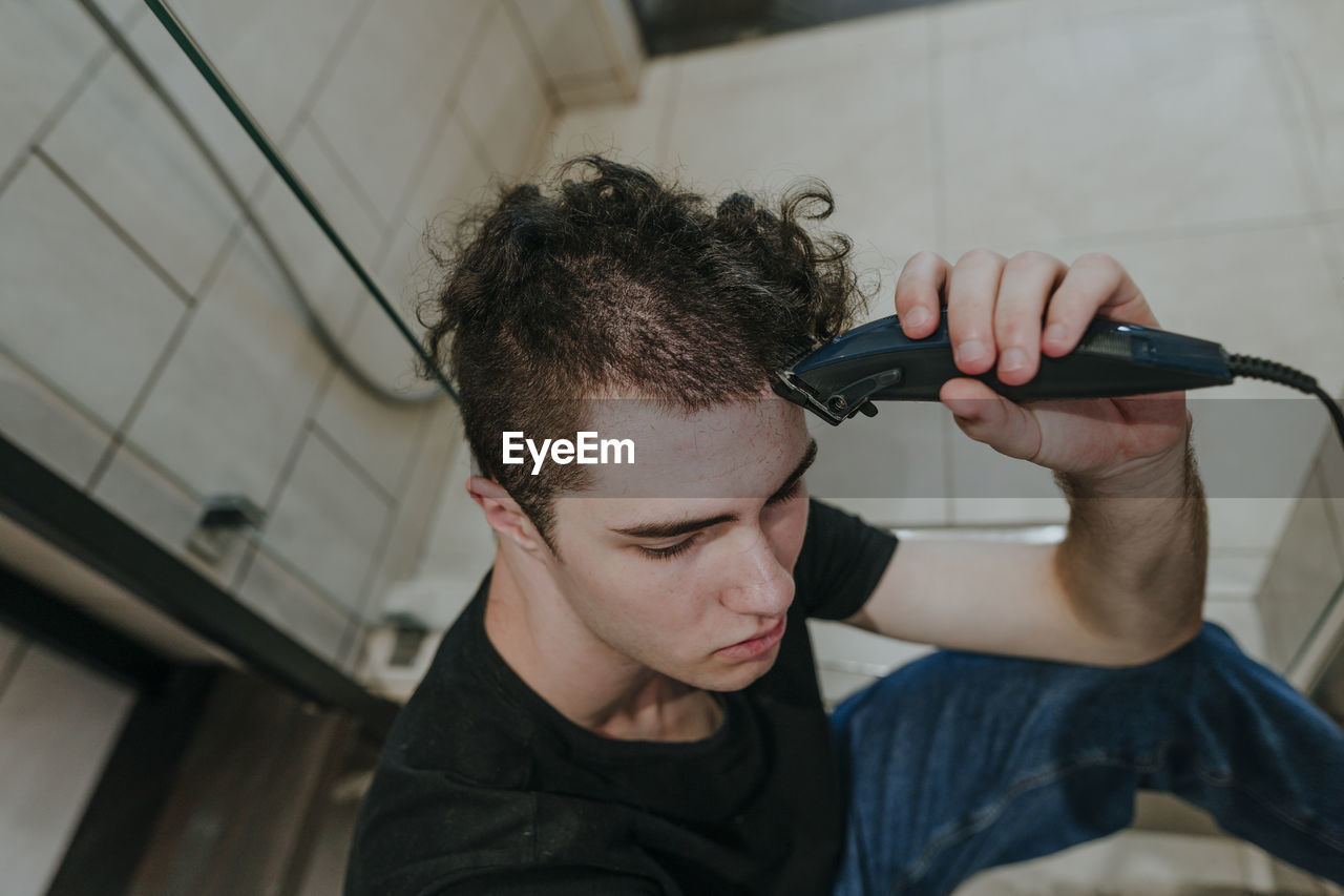 Sad young man with electric razor shaving his own hair in bathroom