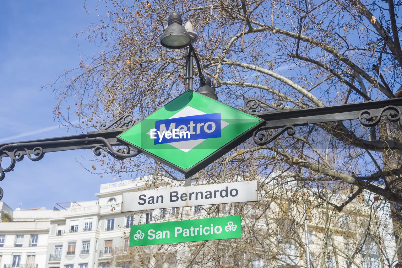 sign, communication, text, tree, road sign, western script, guidance, low angle view, sky, architecture, bare tree, no people, nature, road, day, city, plant, built structure, directional sign, information sign, building exterior, symbol, branch, arrow symbol, outdoors, street, traffic sign, signage, street sign, clear sky, traffic arrow sign, lighting