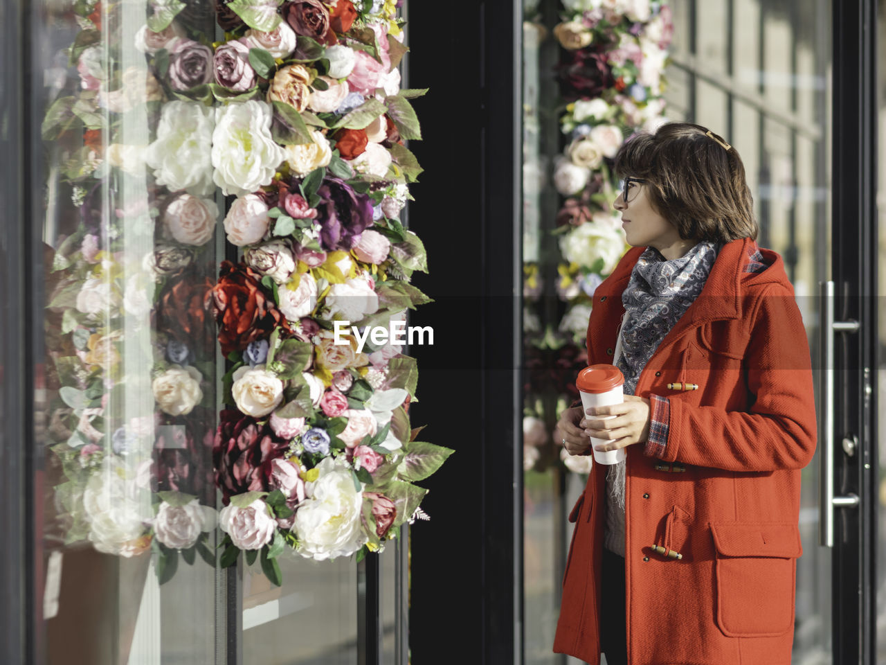 Woman in red duffle coat walks pass florist shop. woman goes along sunny side of street