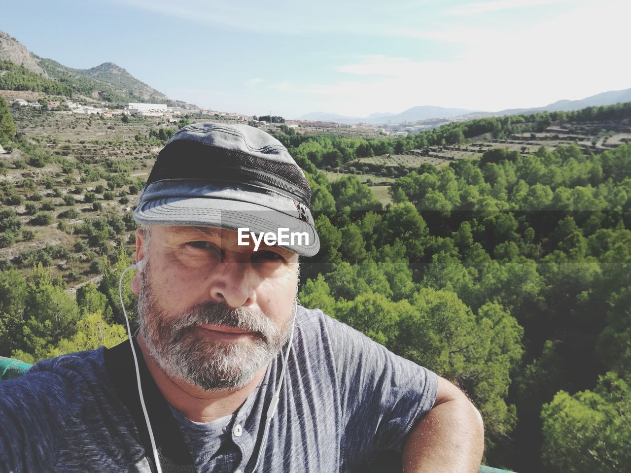 Portrait of man wearing cap listening music against landscape during sunny day