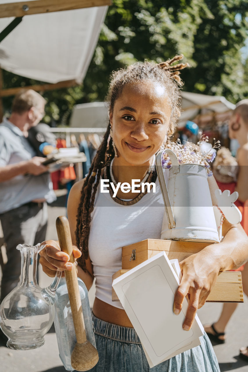 Smiling young woman holding different objects while shopping at flea market