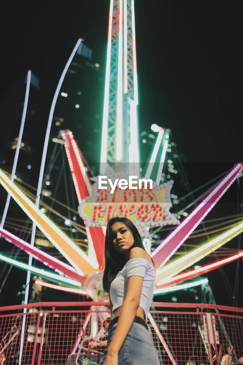 Portrait of young woman standing illuminated amusement park ride at night