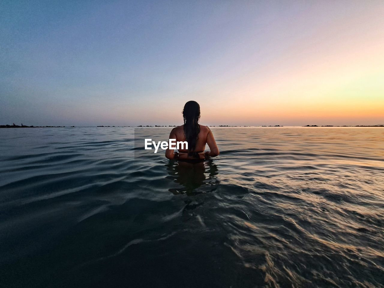 Rear view of girl standing in sea against sky during sunset