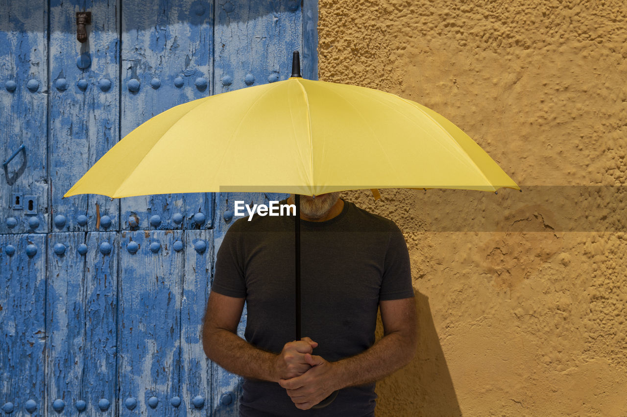 Portrait of adult man with yellow umbrella standing against blue door and yellow wall in summer