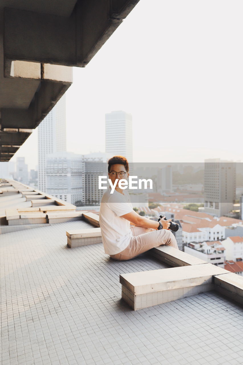 Side view of young man holding digital camera while sitting on floor in city