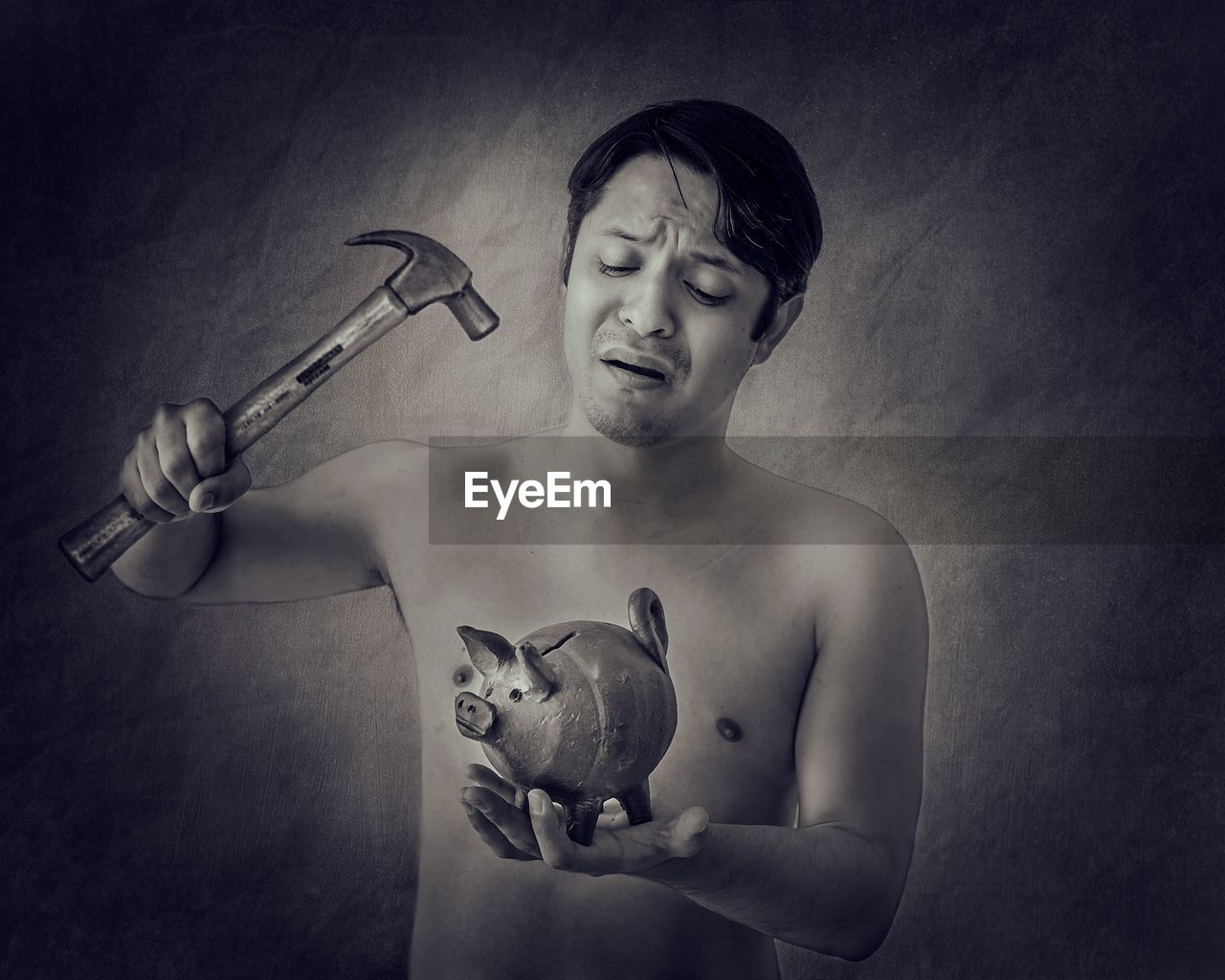 Shirtless mid adult man holding hammer and piggy bank while standing against wall