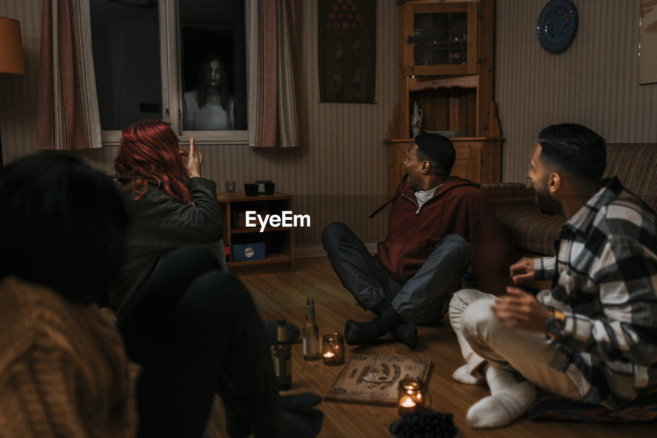 Terrified multiracial friends looking at ghost through window in log cabin