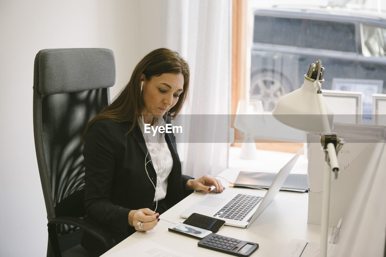 Mature female realtor listening to headphones while looking at laptop in office