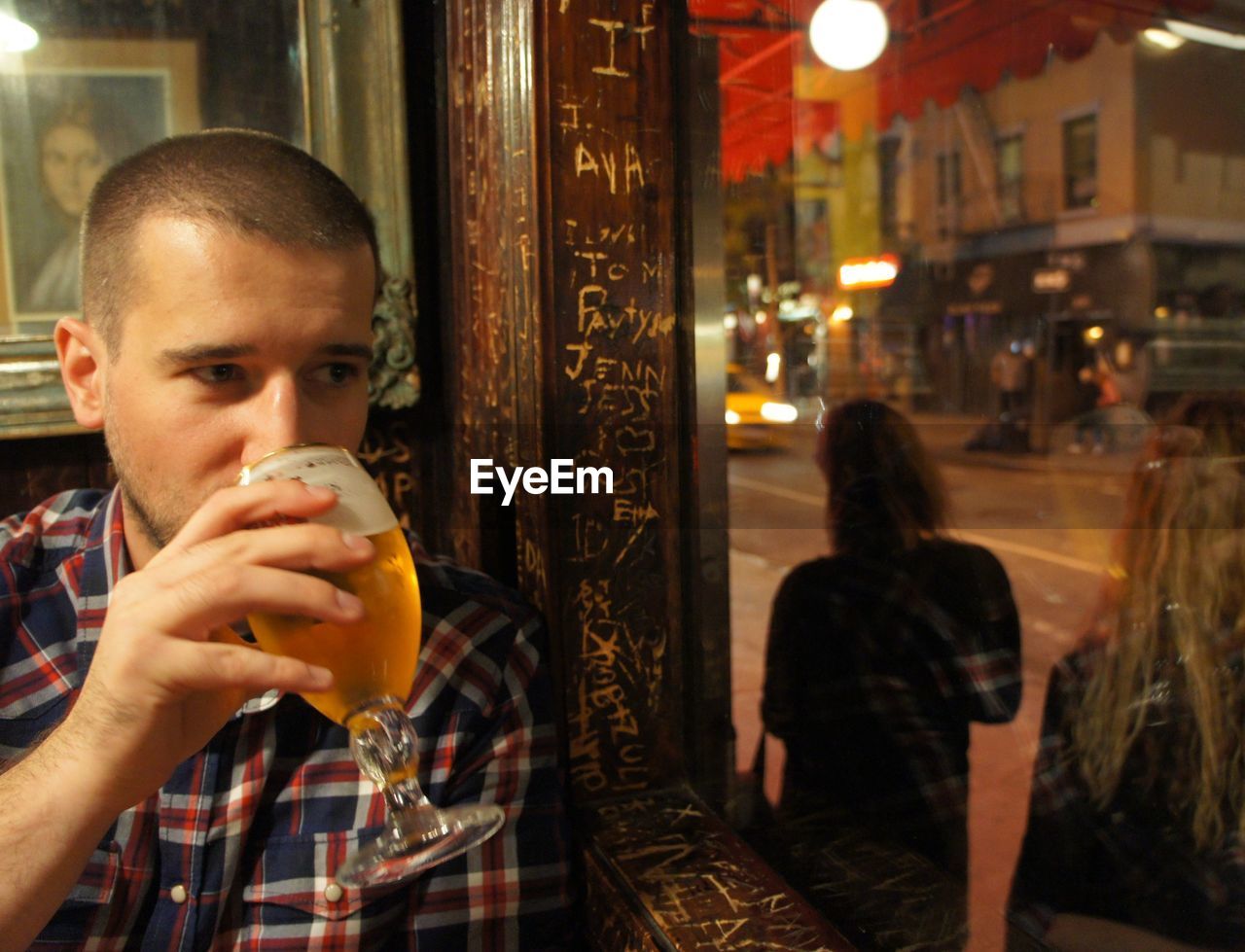 Young man drinking beer while looking out through glass window at restaurant
