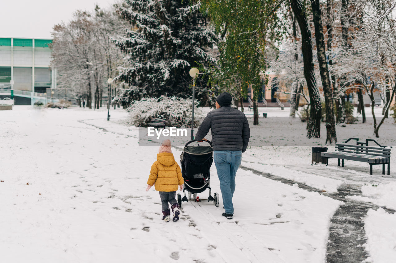 Rear view of father with stroller by daughter walking on snow covered land