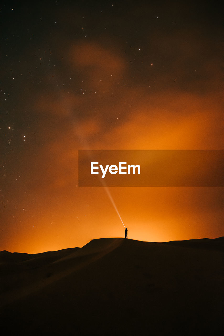 Silhouette person on land against sky during sunset in the desert full of stars