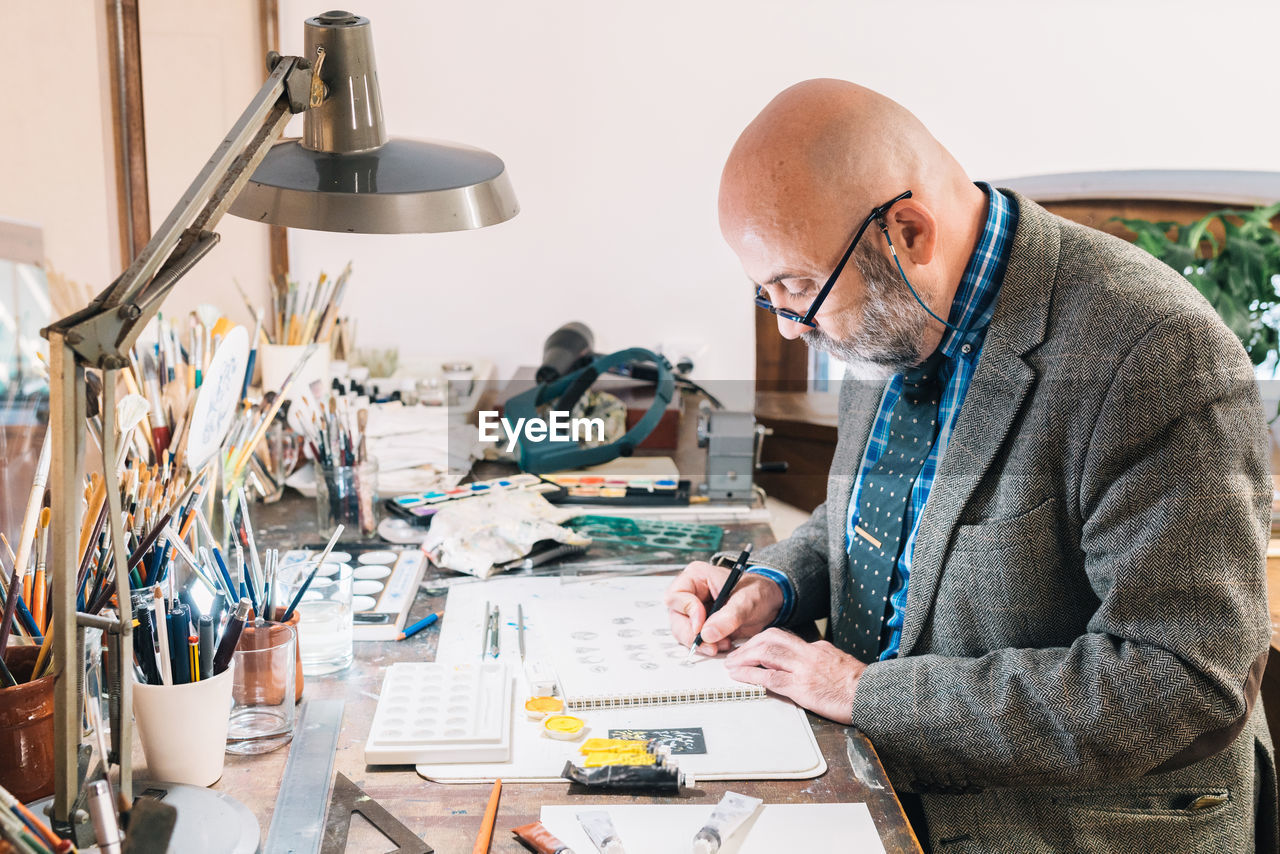 Side view of focused creative mature male designer working on jewelry designs and drawing sketches while sitting at table with professional supplies in workplace