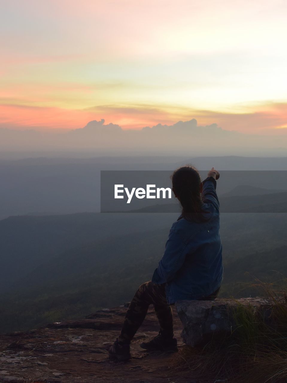 Rear view of woman sitting on mountain against sky at sunset