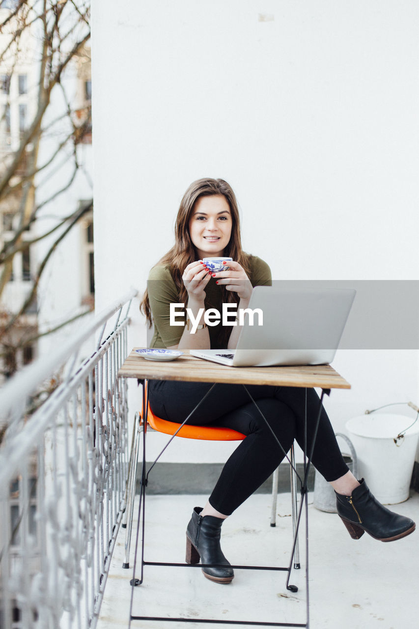 Portrait of smiling young woman with laptop on table having coffee at balcony