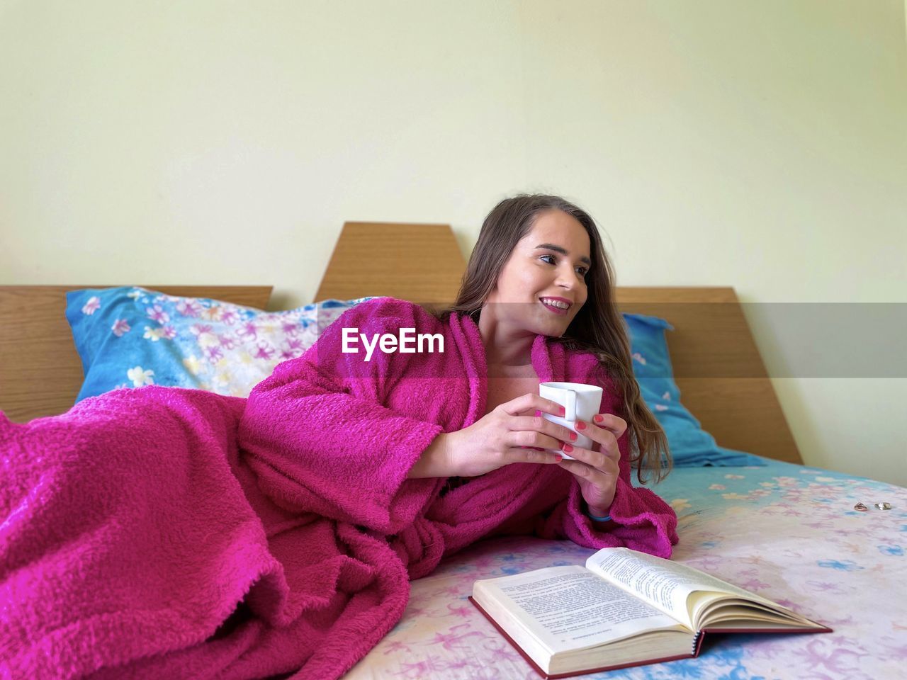 Woman wearing pink bathrobe lying down on the bed holding a cup of coffee in her hands