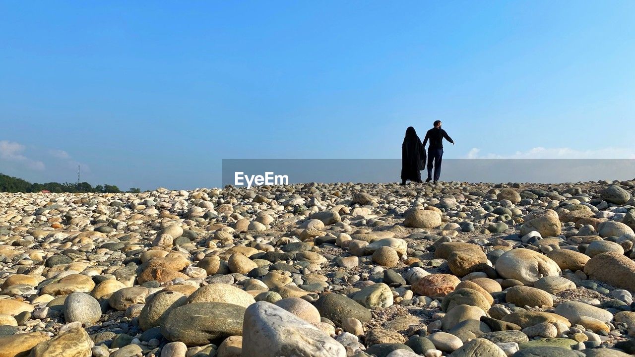 sky, rock, sea, nature, coast, shore, land, stone, men, adult, pebble, two people, rear view, day, standing, full length, leisure activity, copy space, beach, sand, horizon, blue, clear sky, scenics - nature, ocean, beauty in nature, lifestyles, togetherness, women, outdoors, tranquility, sunlight, holiday, water, tranquil scene, travel, environment, trip, vacation, non-urban scene, gravel, sunny, landscape, geology