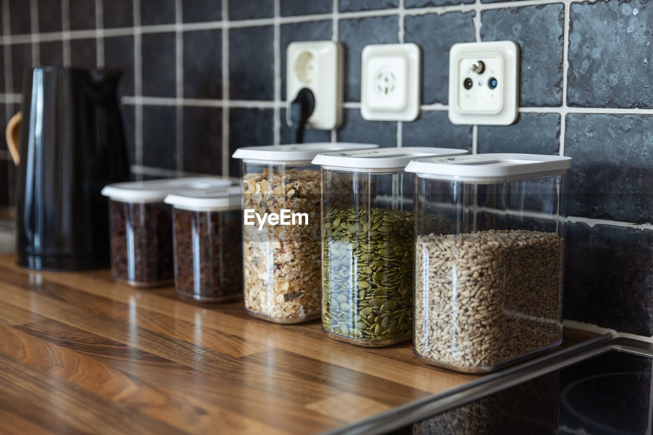 Plastic containers with muesli and sunflower and pumpkin seeds placed near coffee beans and electric kettle on counter in kitchen