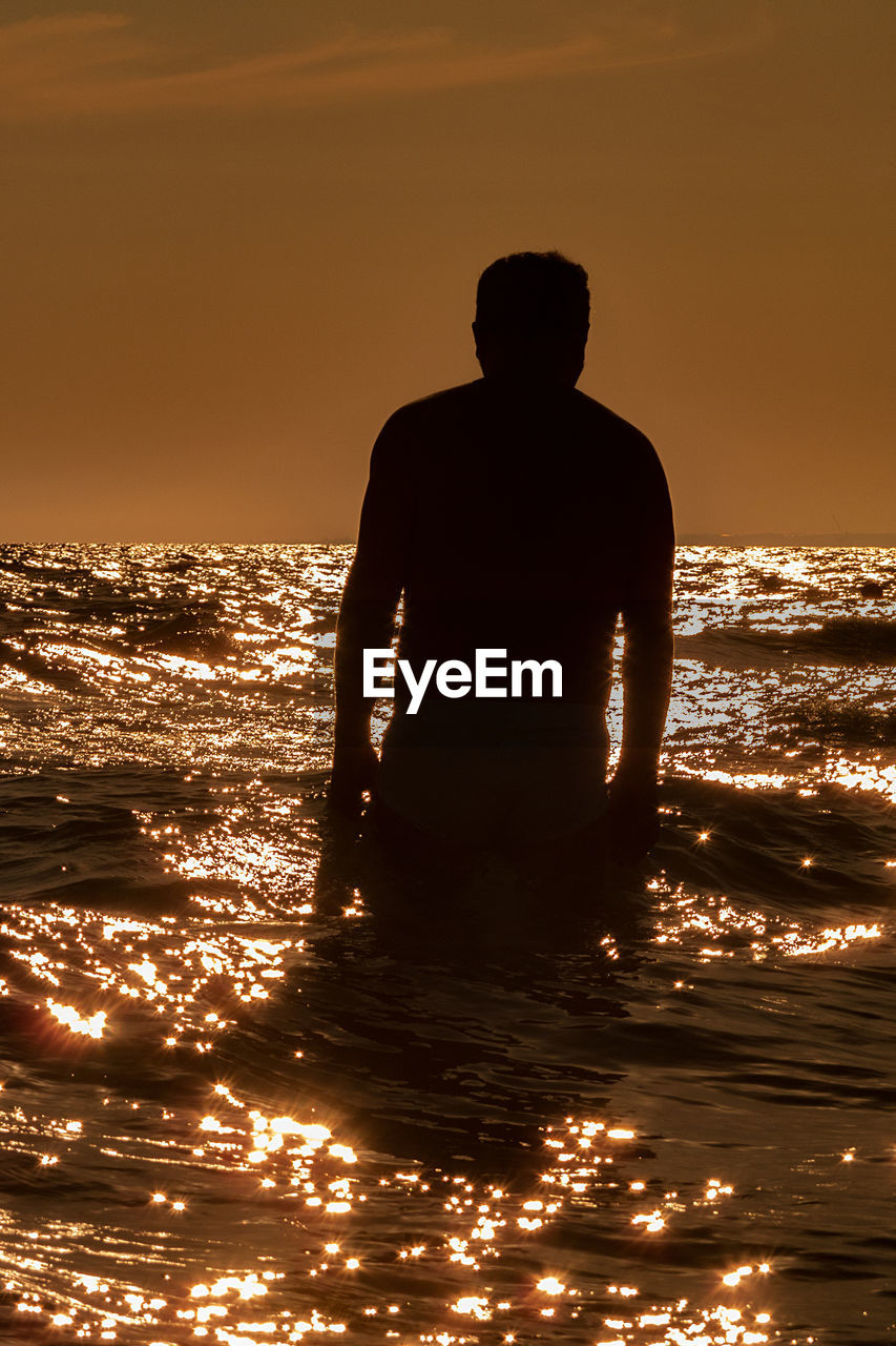 SILHOUETTE MAN STANDING IN SEA AGAINST SKY DURING SUNSET