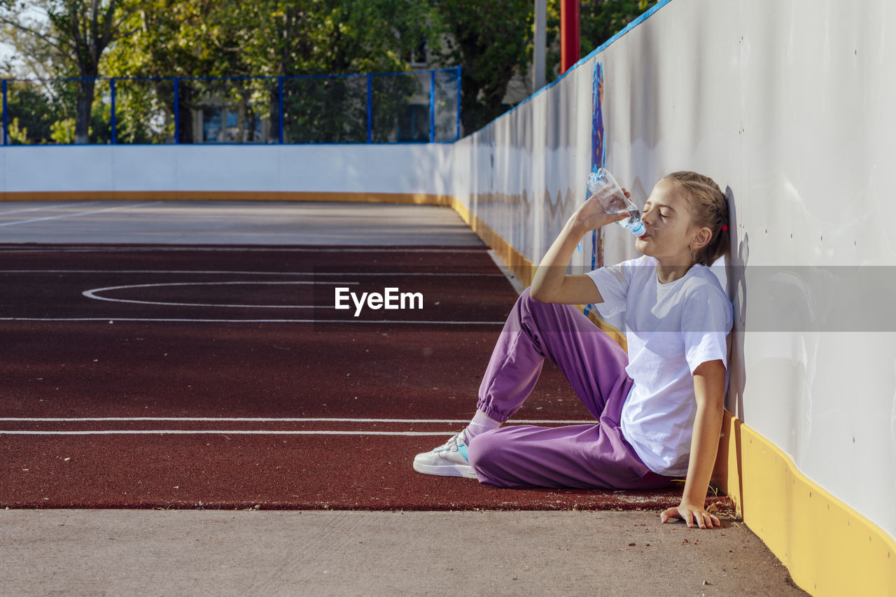 Girl drinks water while sitting on rubber coating on basketball court. sport, active lifestyle