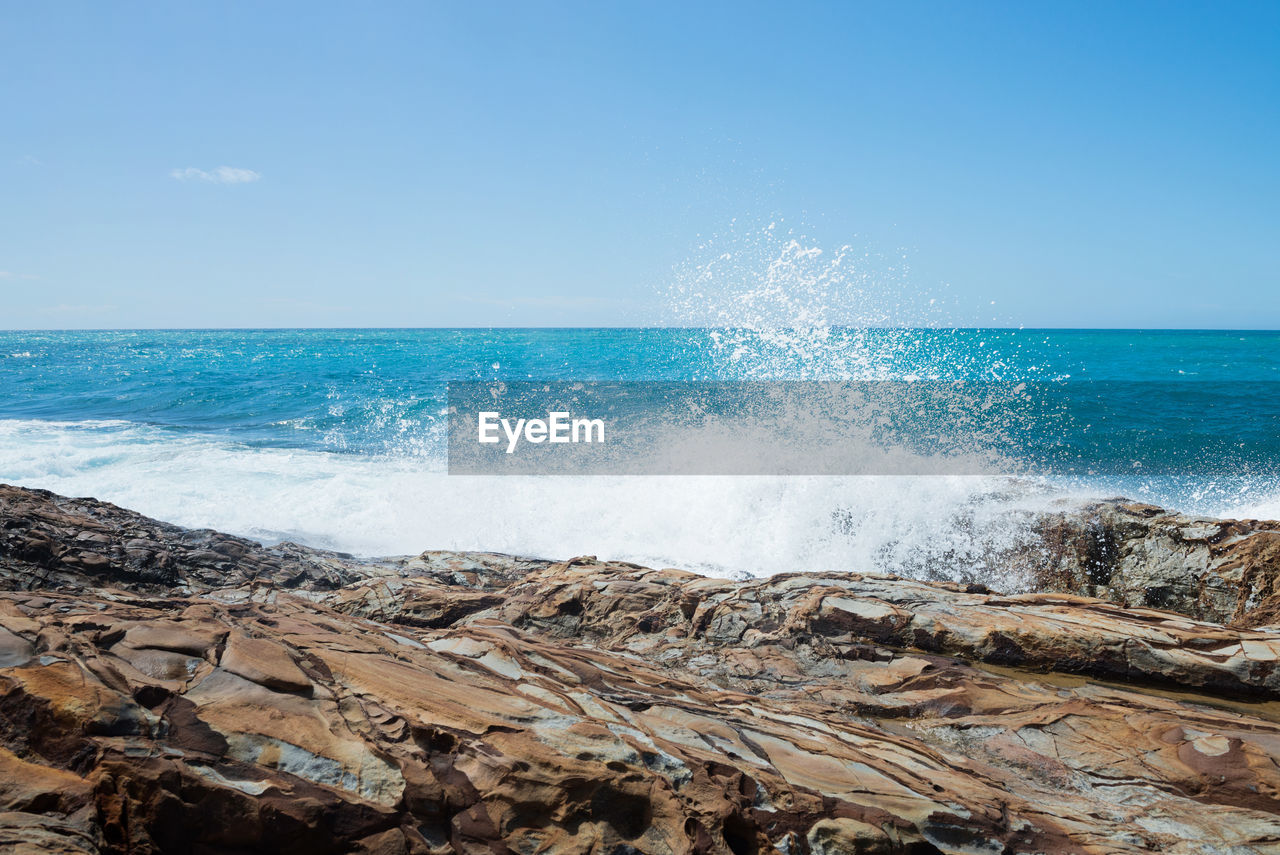 Waves splashing on rocks at shore against blue sky