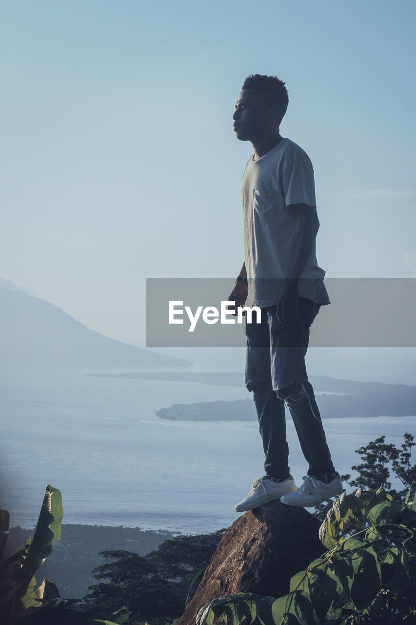 MAN STANDING ON ROCK LOOKING AT SEA SHORE AGAINST SKY