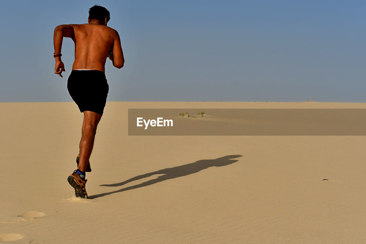 Full length of shirtless young man running on sand at desert during sunny day