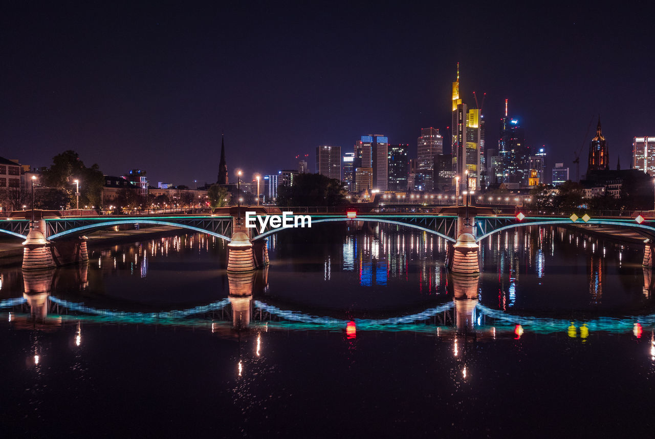 Illuminated bridge over river by buildings against sky at night