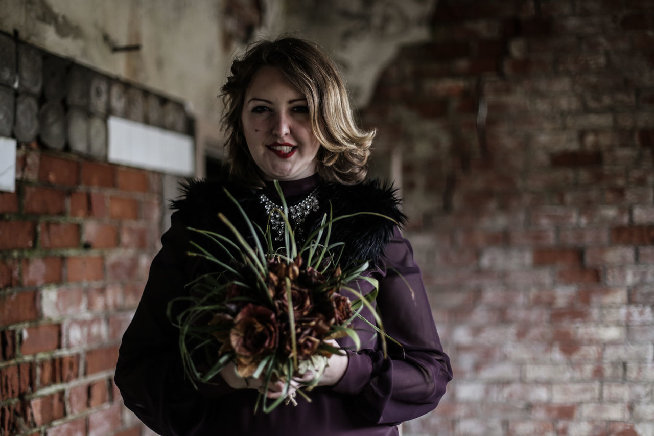 Portrait of young woman holding dried flowers against wall