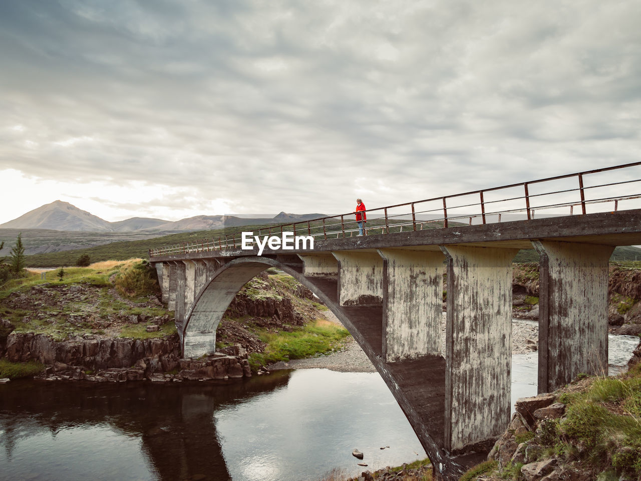 Woman is standing on old bridge over river against cloudy sky