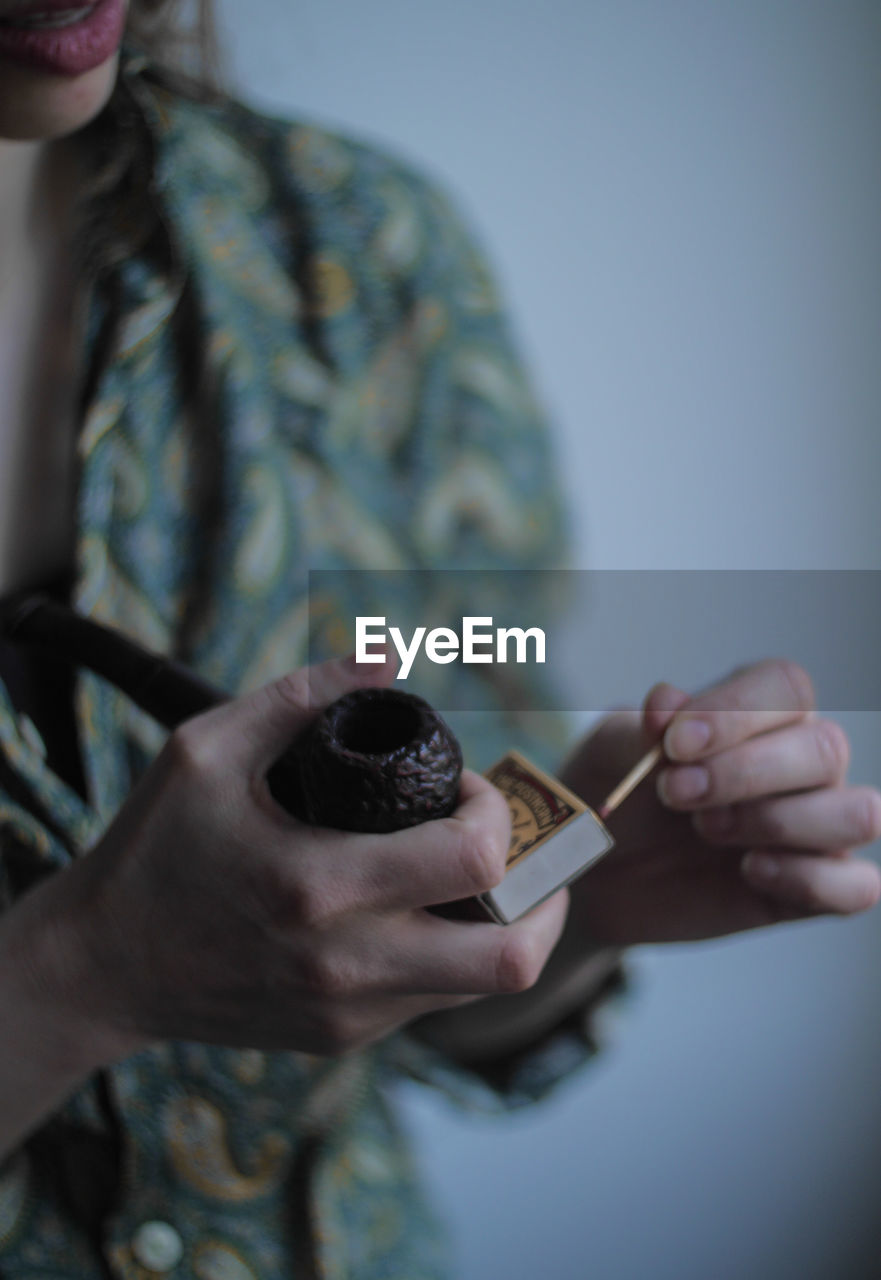 Midsection of mid adult woman holding matchstick and pipe while standing against wall