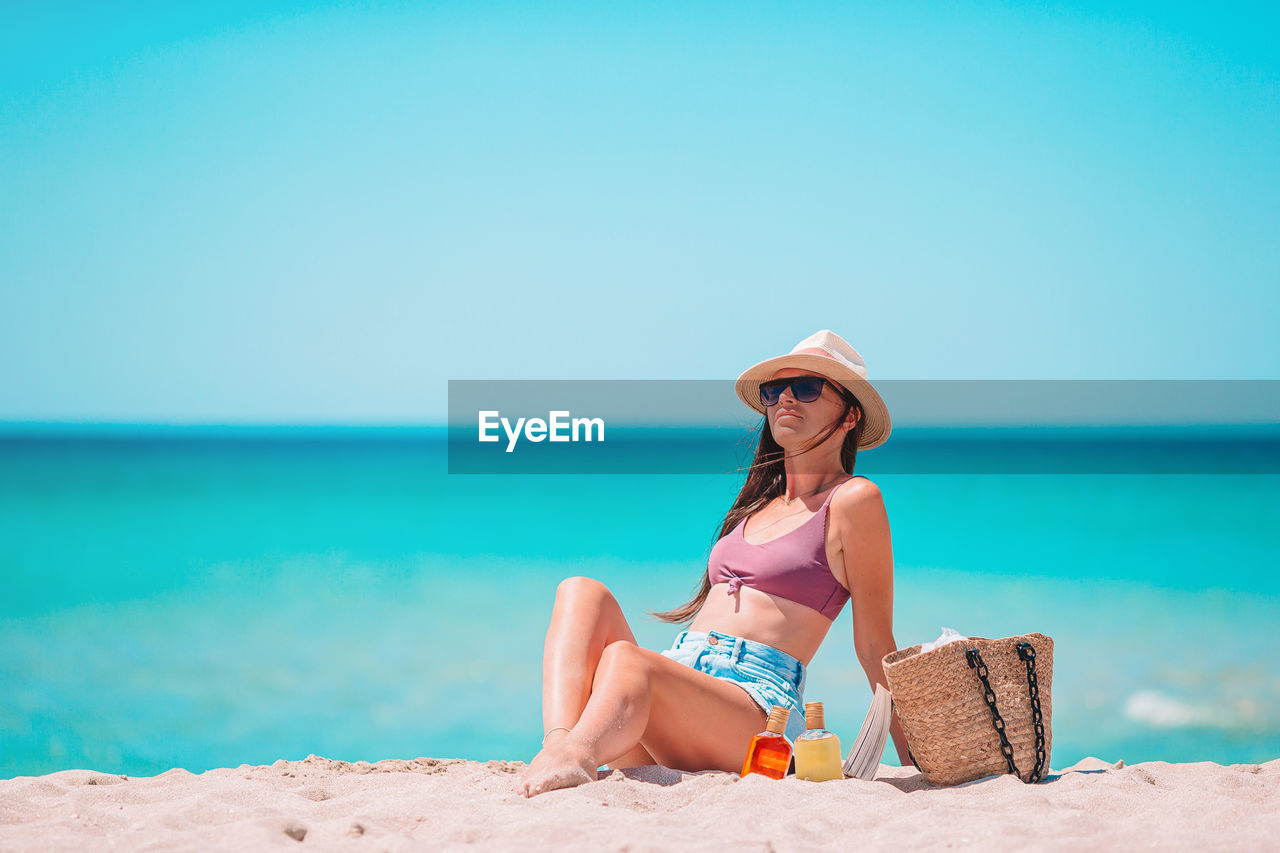 YOUNG WOMAN SITTING ON BEACH AGAINST SEA