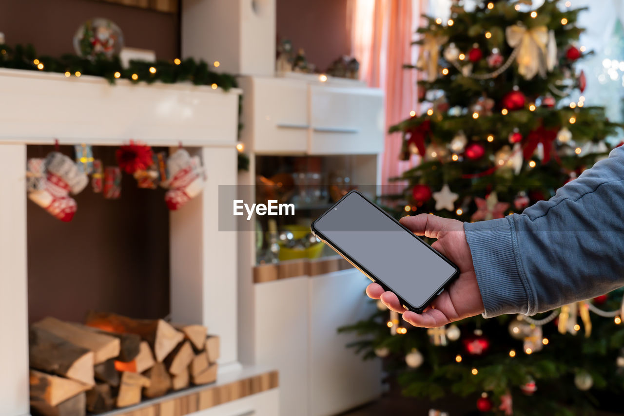 A male hand holds a smartphone. the man standing in front of christmas tree and fireplace console.