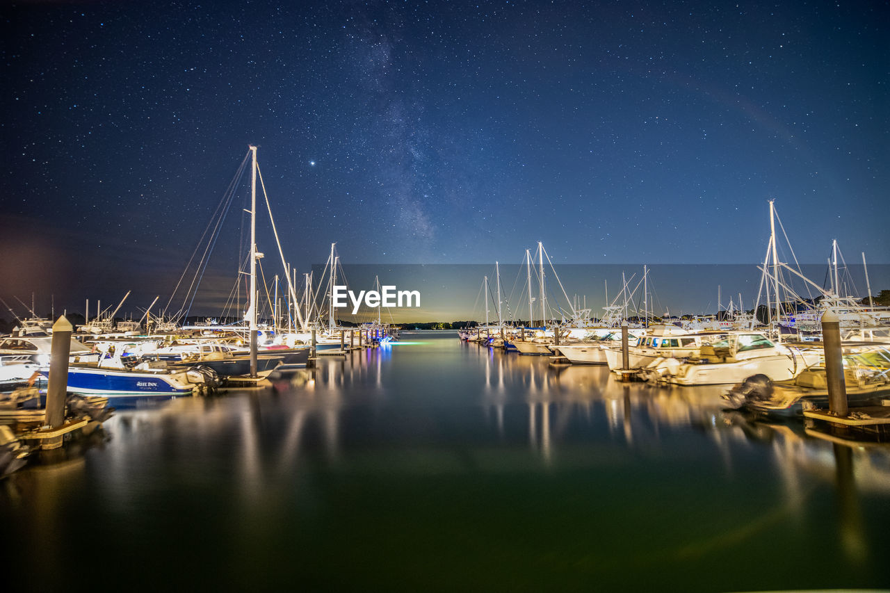 Sail boats docked in the harbor reflecting under the milky way sky.