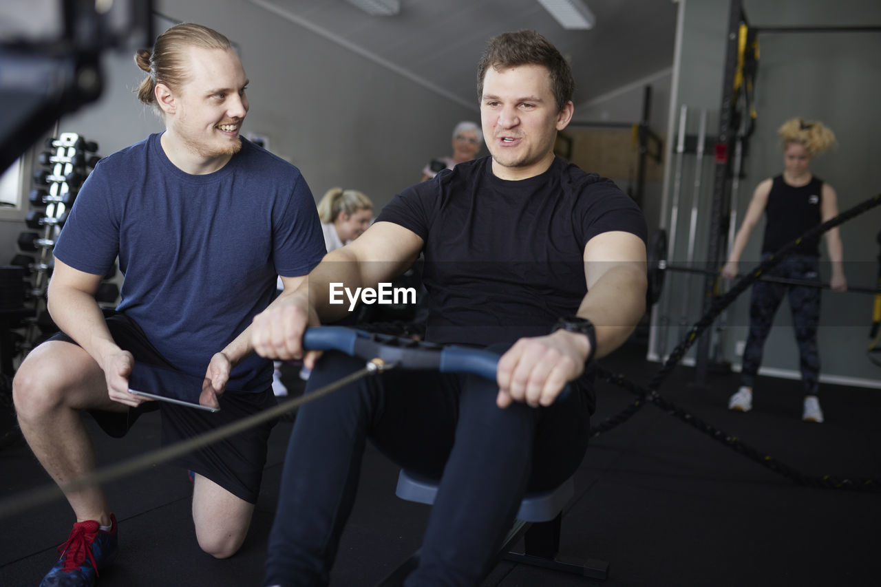 Smiling male instructor encouraging man exercising on rowing machine at gym