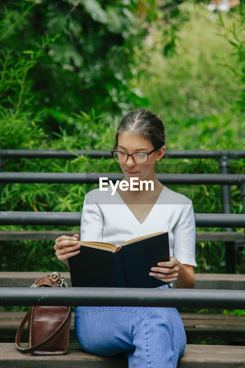 Young woman wearing eyeglasses reading book while sitting on bench in park