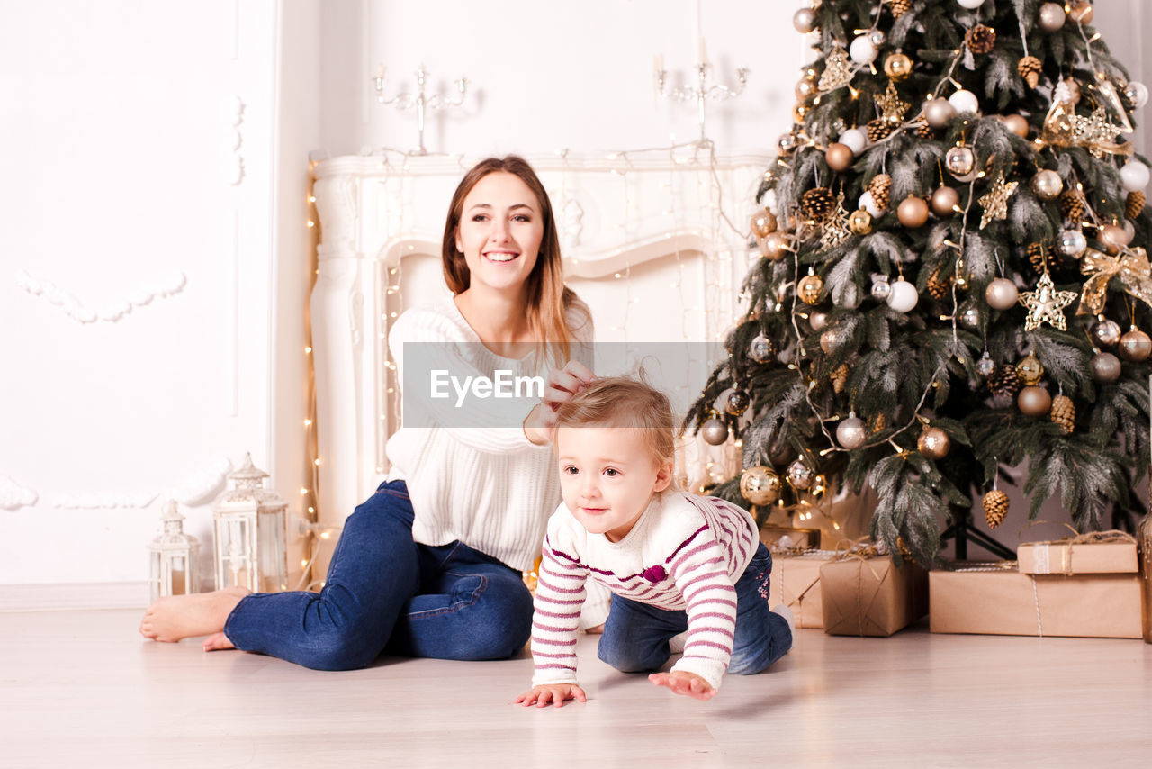 child, childhood, family, christmas tree, emotion, women, parent, baby, happiness, female, smiling, indoors, togetherness, adult, tree, toddler, positive emotion, christmas, two people, one parent, person, men, home interior, celebration, full length, bonding, lifestyles, holiday, plant, domestic life, sitting, love, cute, portrait, flooring, cheerful, nature, domestic room, portrait photography, blond hair, living room, father, decoration, casual clothing, looking at camera, clothing, innocence, joy, christmas decoration
