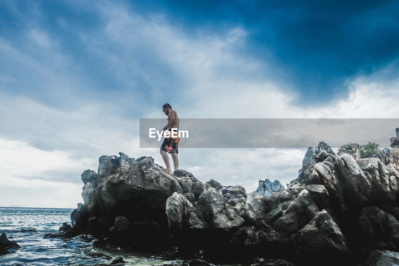 Low angle view of shirtless man standing on rock by sea against cloudy sky