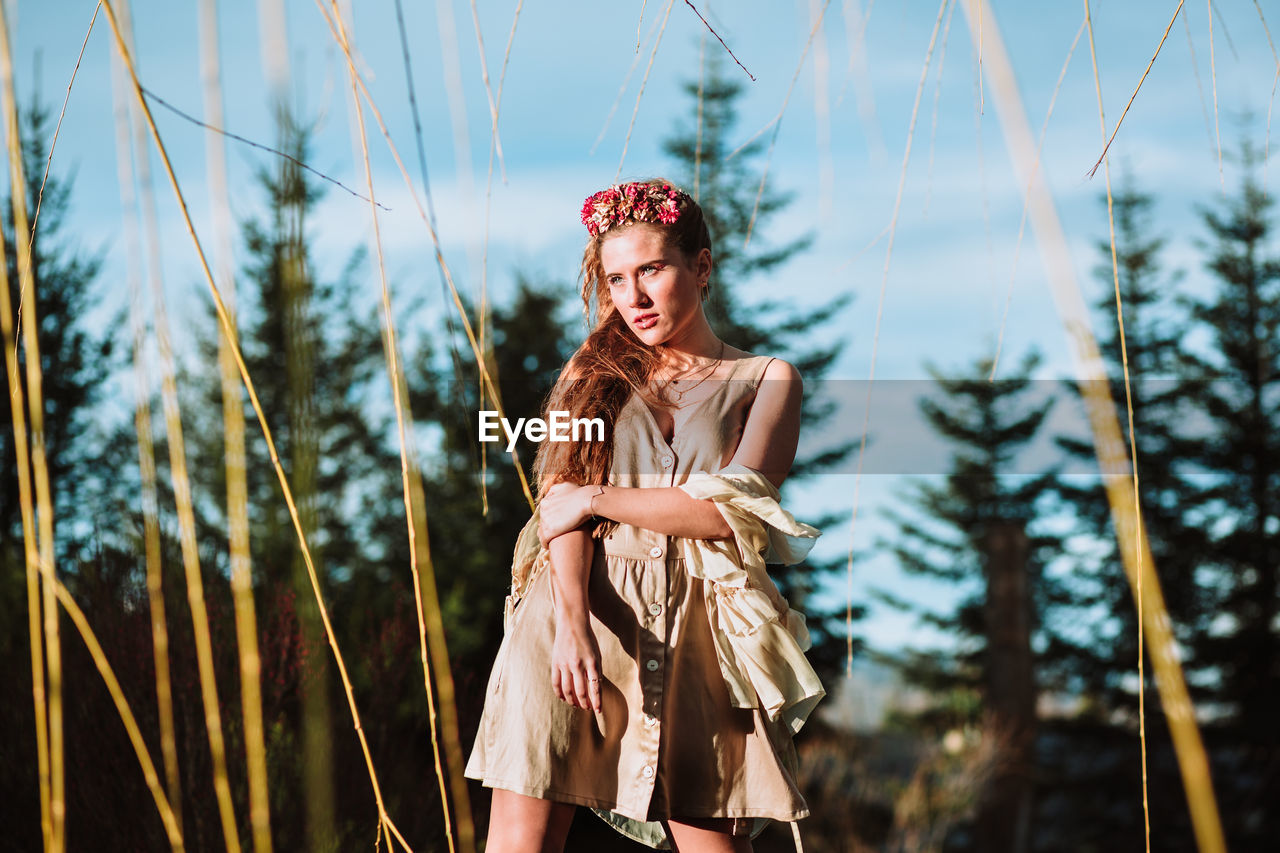 Full body of stylish young long haired female in trendy dress standing on meadow with leafless trees against mountains