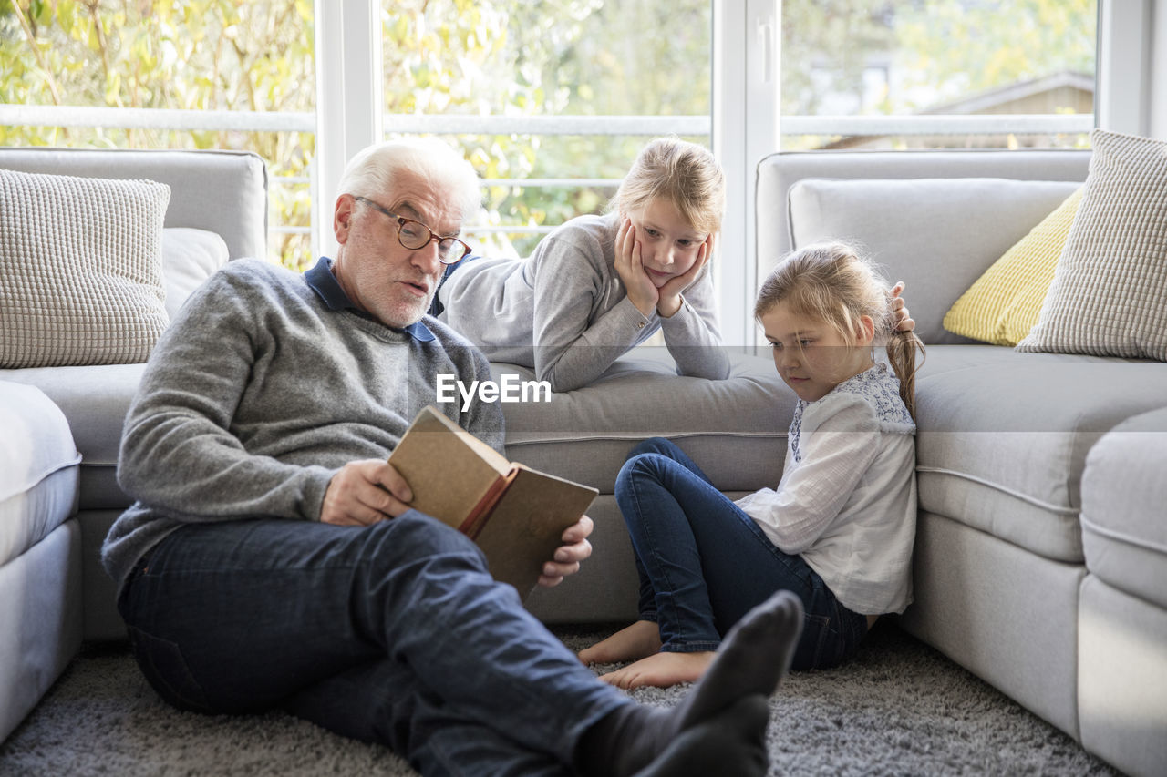 Two girls and grandfather reading book in living room