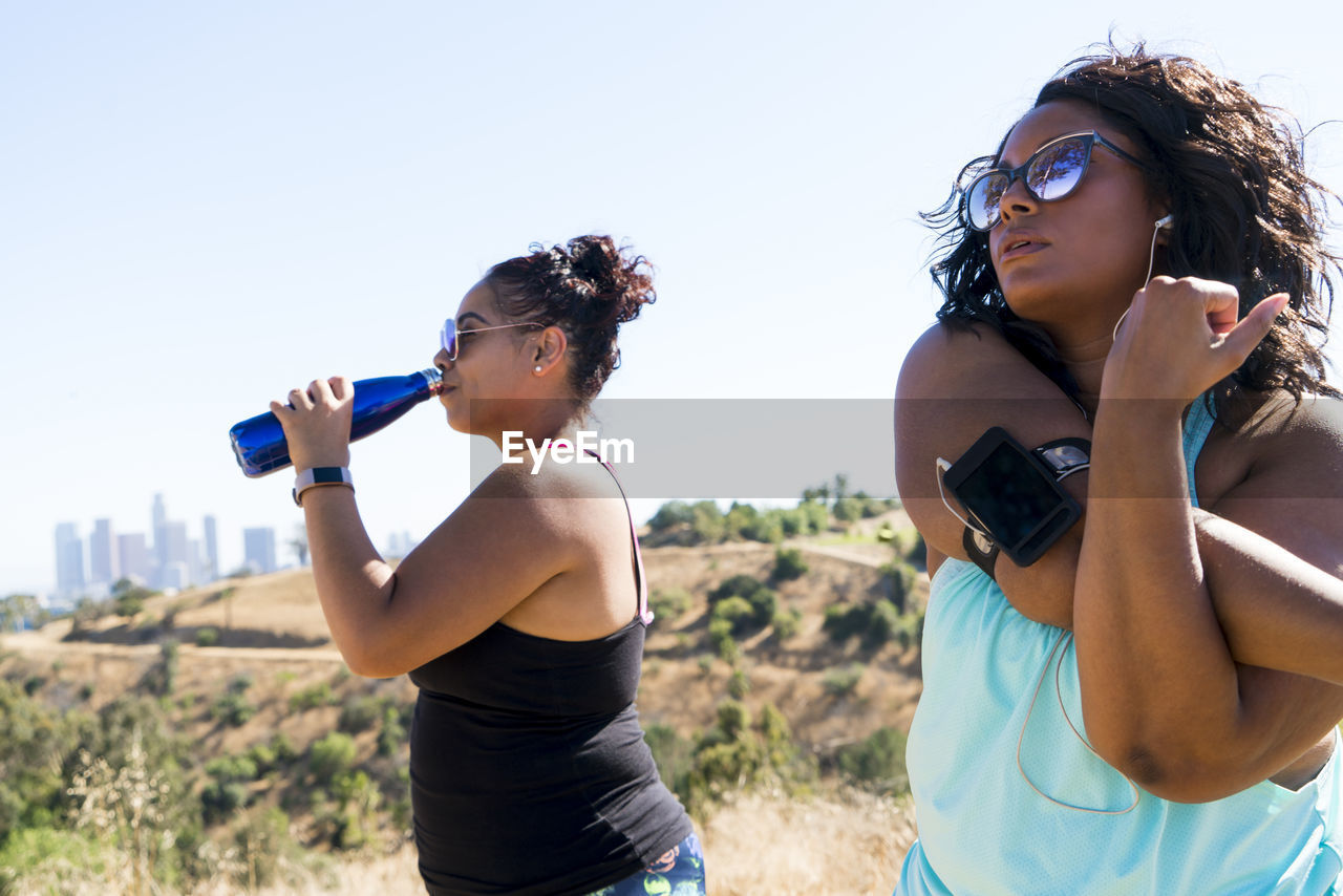 Woman stretching arms while female friend drinking water against clear sky