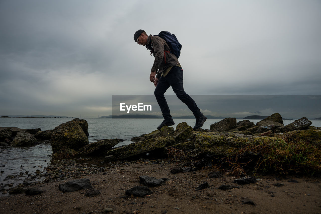 Hiker walks across mossy muddy stones from structure on bayside beach