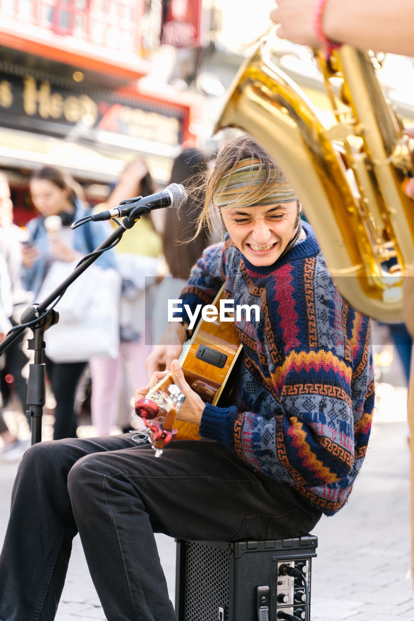 Vertical photo of a young man singing while playing guitar next to a saxophonist in the street