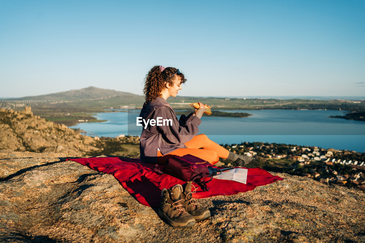 Side view of happy young female hiker in casual clothes eating sandwich while sitting on blanket on rocky hill over sea against cloudless blue sky