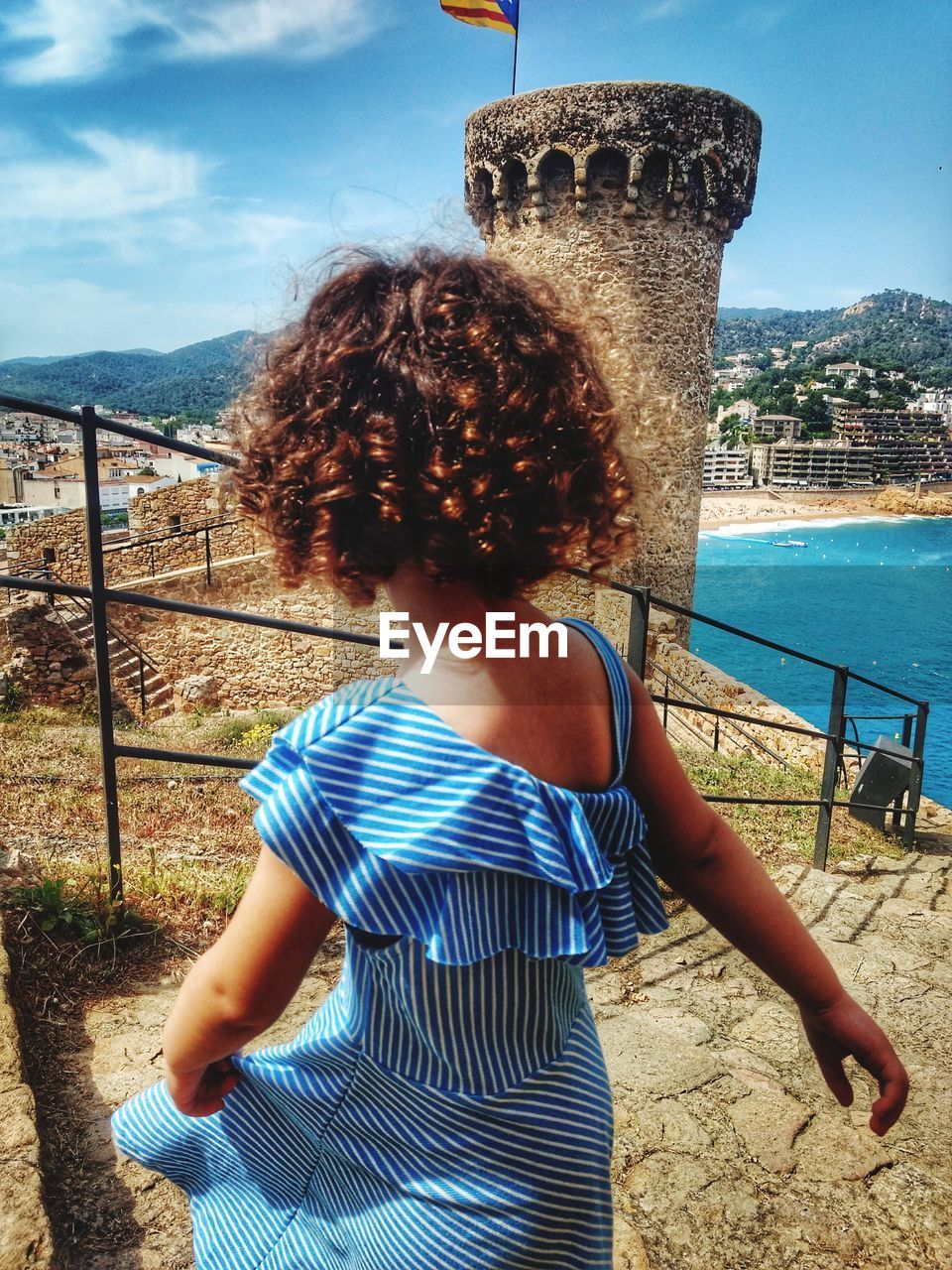 Rear view of girl with curly hair standing on steps against blue sky during sunny day