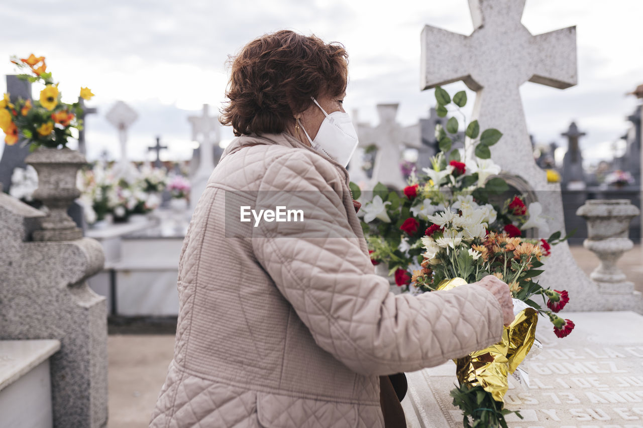 Senior woman wearing protective face mask putting flowers on tombstone in cemetery